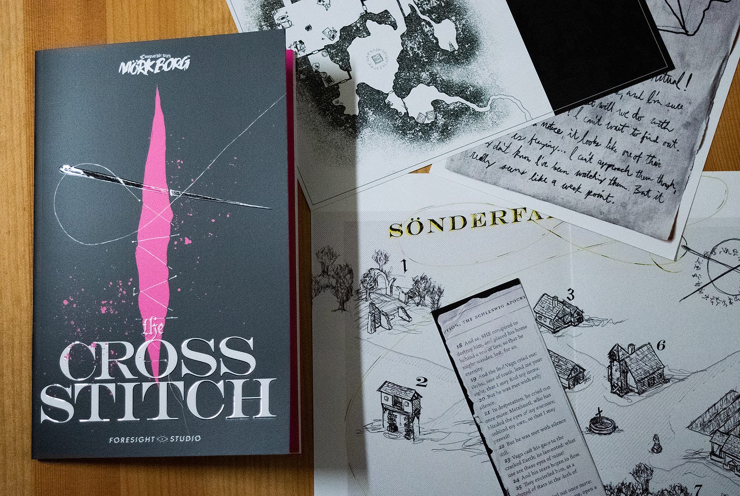 A photograph of a zine on a table, closed with the cover facing up on the left side of the image. The cover is black with a magenta wound going from top to bottom, and a white needle suturing the wound shut. At the bottom of the cover are the words "the Cross Stitch" in white. On the right side of the image, there are several scattered maps and notes that come packaged with the zine.
