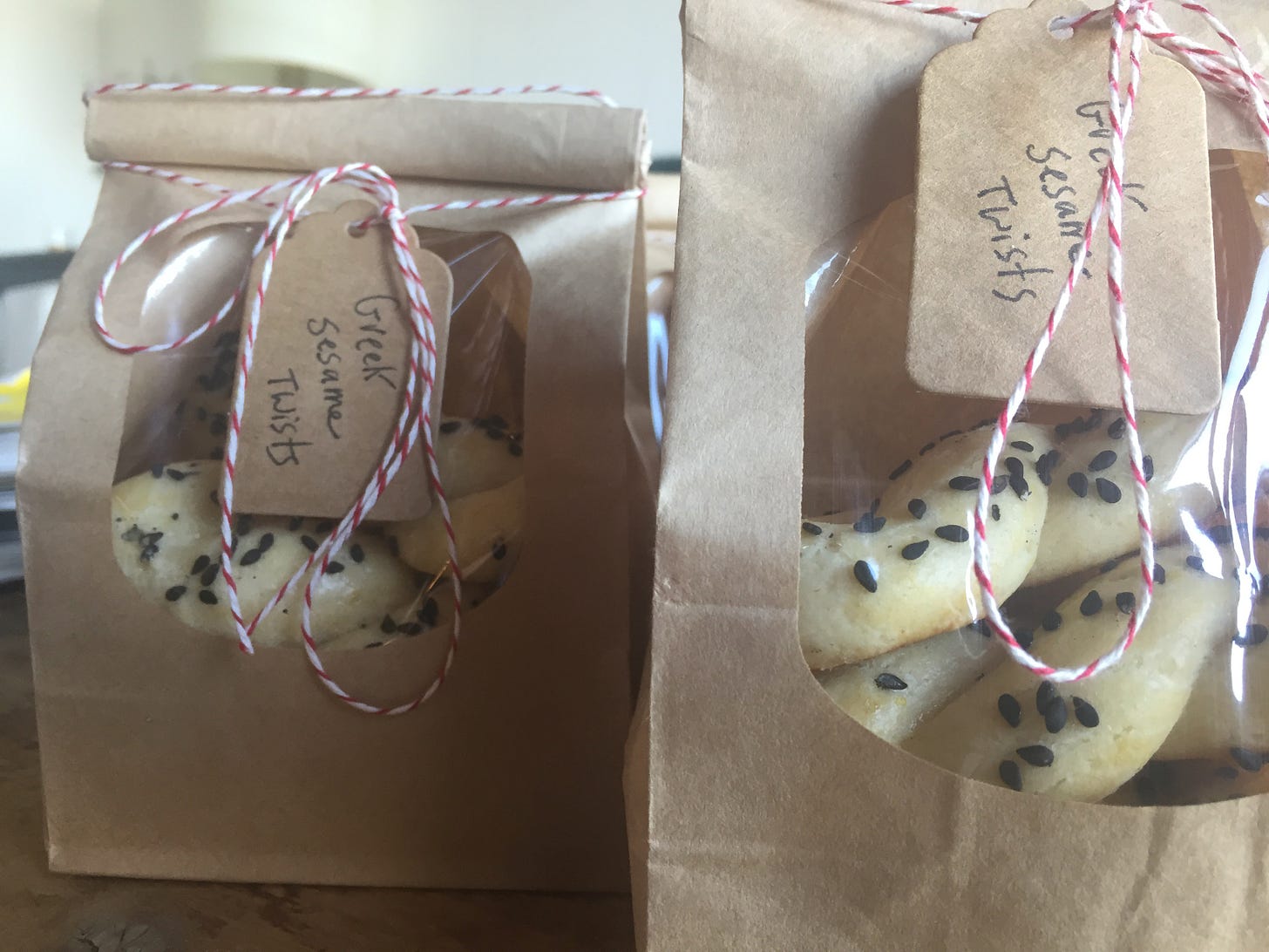 Two small brown bags with windows showing pale cookies studded with sesame seeds inside. The bags are tied around the tops with red and white twine and have small handwritten tags that say ‘Greek Sesame Twists’.