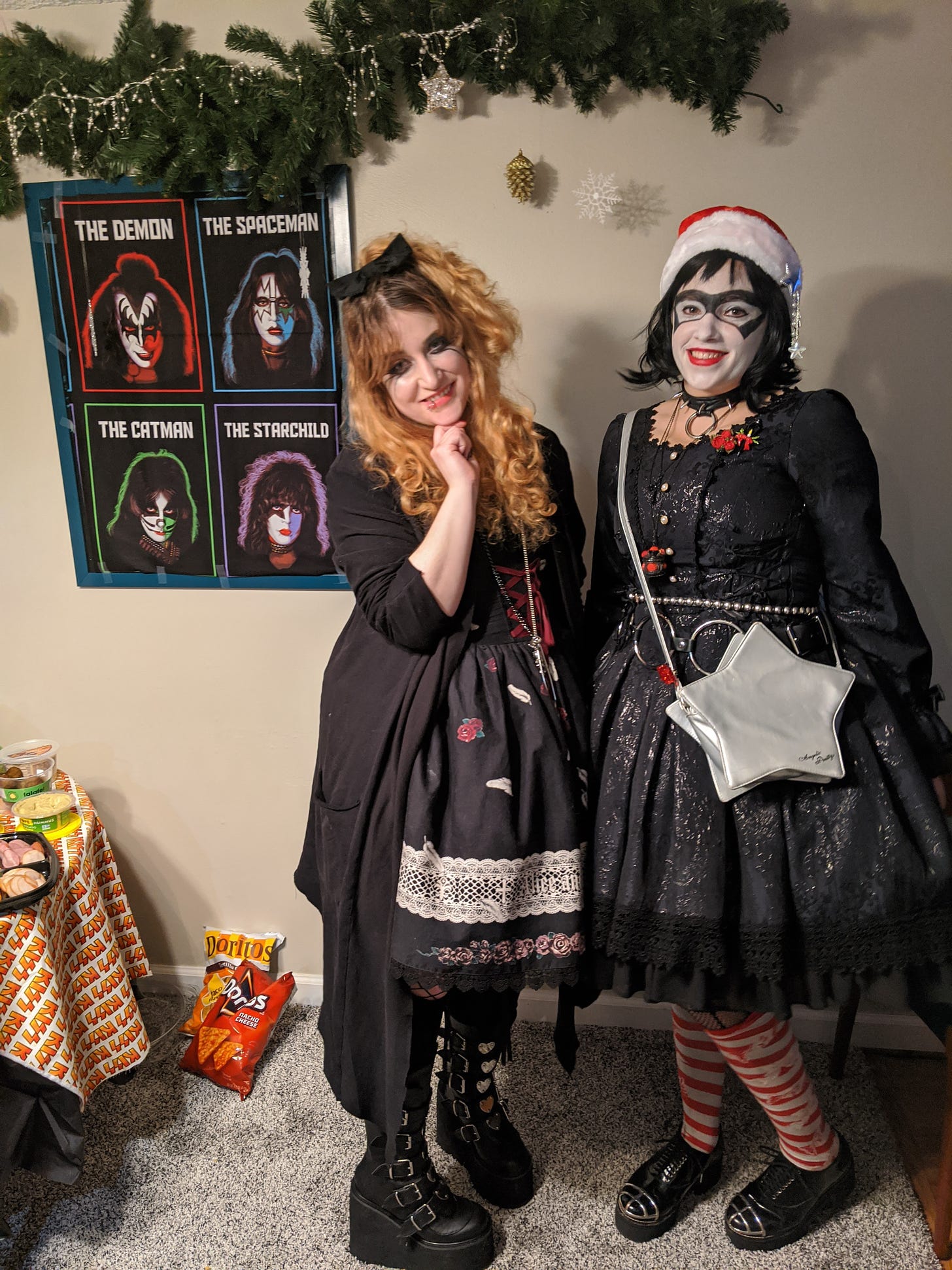 Nif and Lexi (two white women) pose together in Lolita fashion. Nif has mostly blonde hair up in a flirty half-ponytail with a black bow. She is wearing whiskered eye makeup and pale red lipstick. She is wearing a gothic Lolita/rokku gyaru-inspired look with a long cardigan, an AatP dress, and Demonia boots. Lexi has black hair and is wearing a santa hat with a silver star clip. She is wearing "The Bandit" KISS makeup. She has a spike collar and big chain belt, red accessories, a silver purse, and she is wearing a damask-print dress by Lief. They are standing next to a KISS poster and some Christmas decorations.