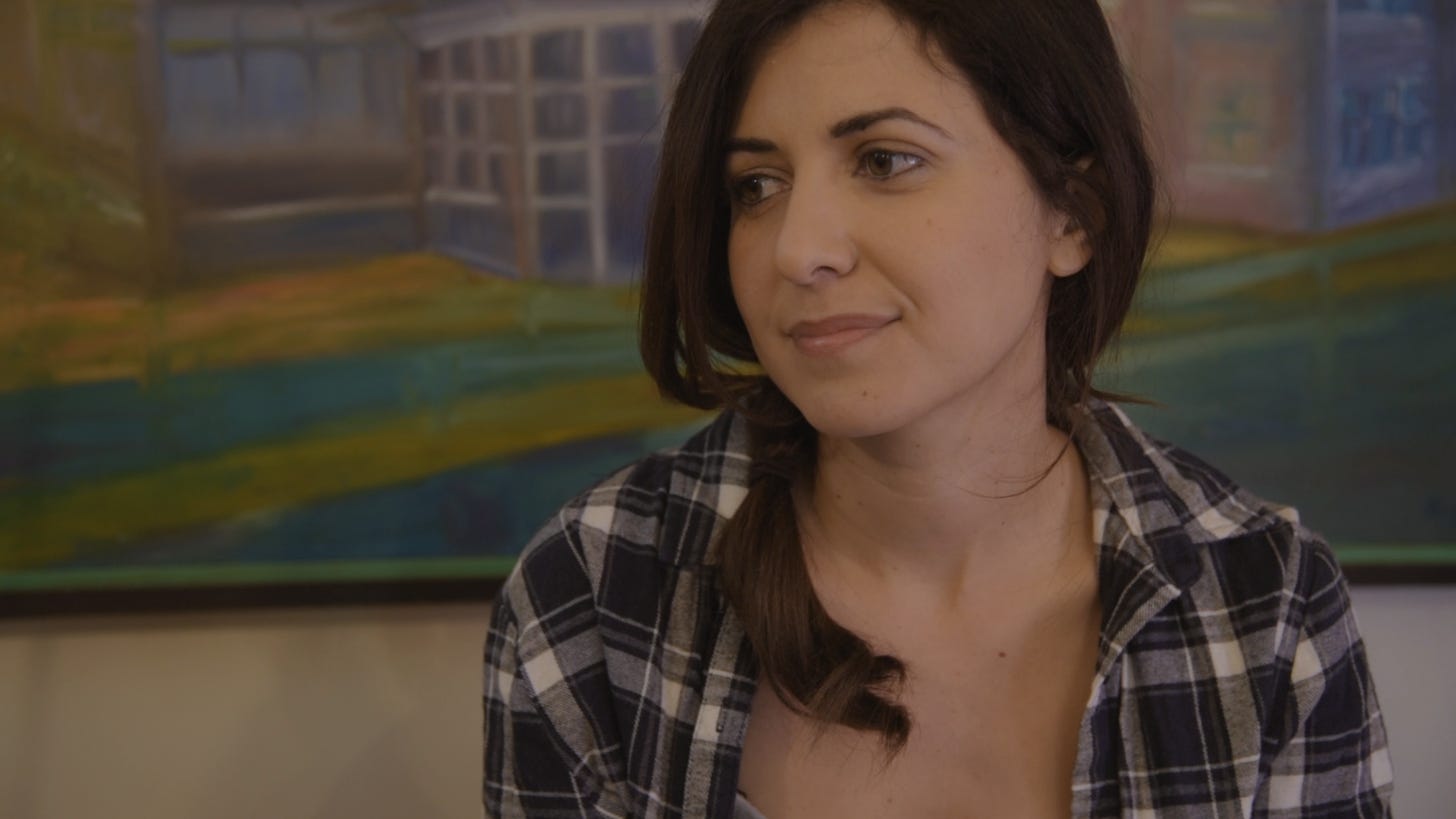 A young Latina woman in a plaid shirt is sitting thoughtfully, gazing to the side, with a background of a large painting behind her. Kristina Pupo is the actress playing Ana in the YouTube series 'Rational Creatures' based on Jane Austen's 'Persuasion.'