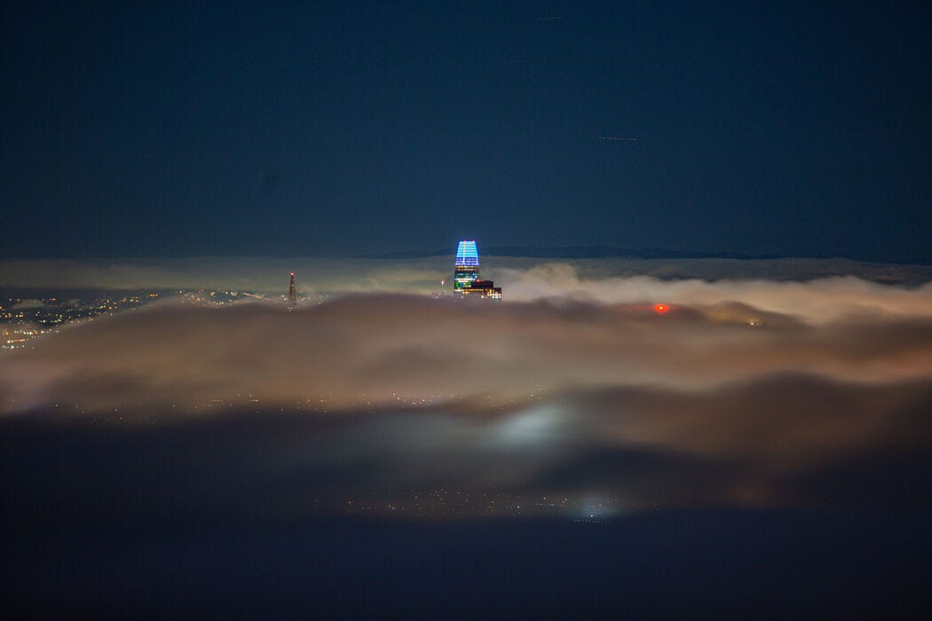 A distant view of a few San Francisco skyscrapers barely poking through a layer of dense fog at dusk. The fog is illuminated in parts by city lights underneath.