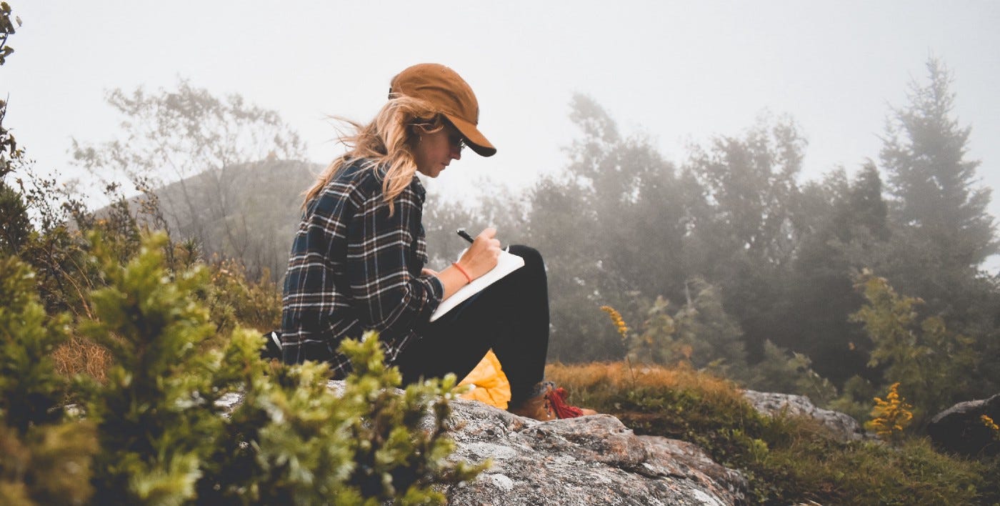 Blonde woman with a brown cap journaling amid green scenery