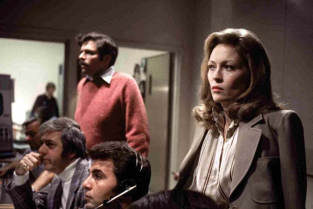 Diane (Faye Dunaway), observes from a broadcasting editing booth. She is wearing a sharp beige suit and striped blouse, and has a taut expression.