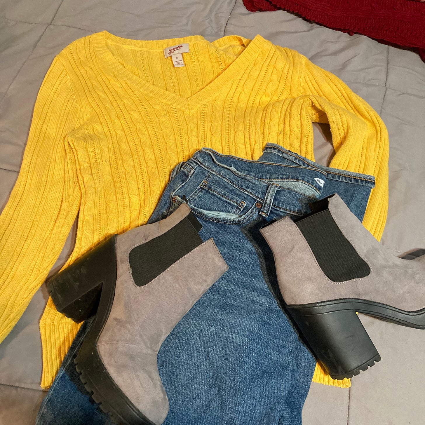 Description: A yellow V-neck sweater, topped by a folded pair of blue jeans, topped by a pair of gray suede platform boots laying on their side are spread out on a gray quilted bedspread.