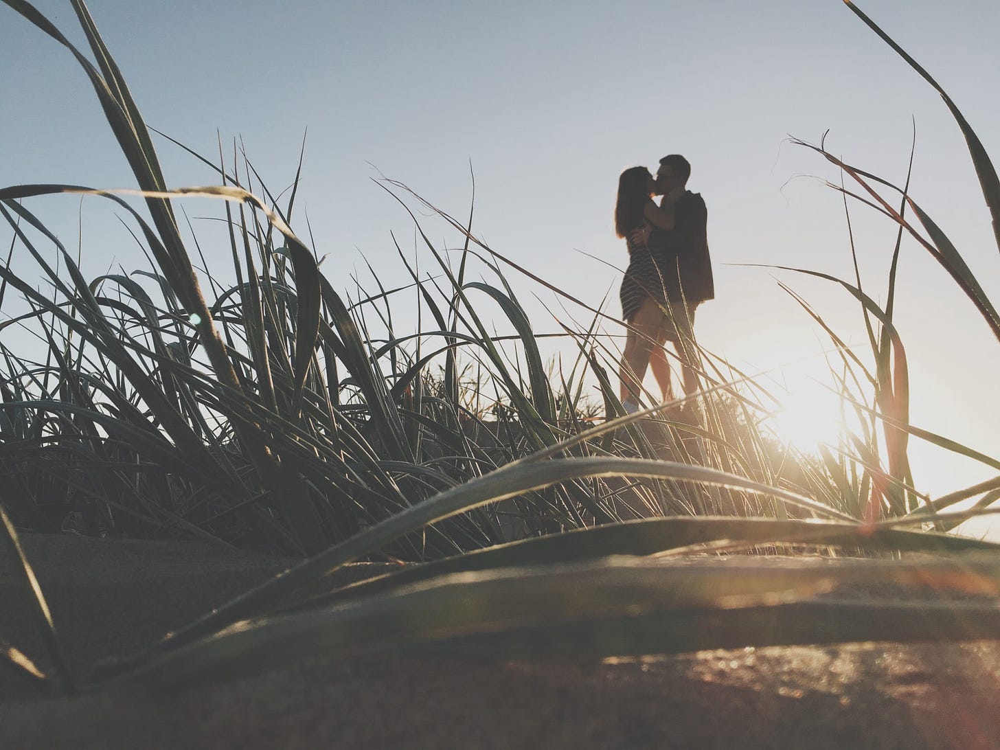 The profile of a couple embraces and kisses as the sun sets over a grassy field.
