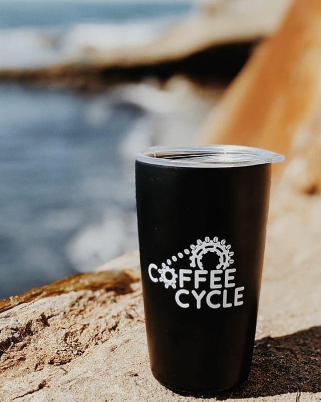 A tall black metal tumbler with lid. On the side it says Coffee Cycle with a bicycle gear. It is resting on a beige, reddish rock at the beach with the ocean blurry in the background.
