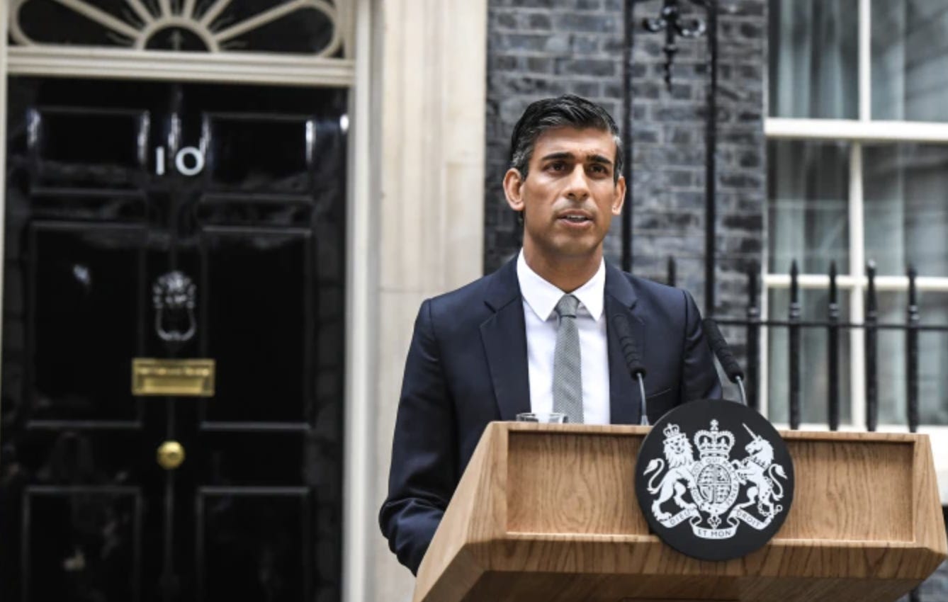 A color picture of an Indian man standing in a dark suit and white shirt outside a grey brick building with “10” on the black door. He stands in front of a brown wooden lectern which has the UK coat of arms on it in white on a black background. It is daytime. The man has short black hair that is starting to grey at the temples. He is in the midst of speaking and is wearing a serious expression.