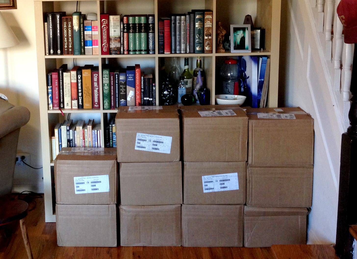 Eleven unopened cardboard cartons sit stacked before a bookcase crammed with books and knickknacks. A lamp and chair are partially visible to the left, with a rising staircase partially visible to the right.