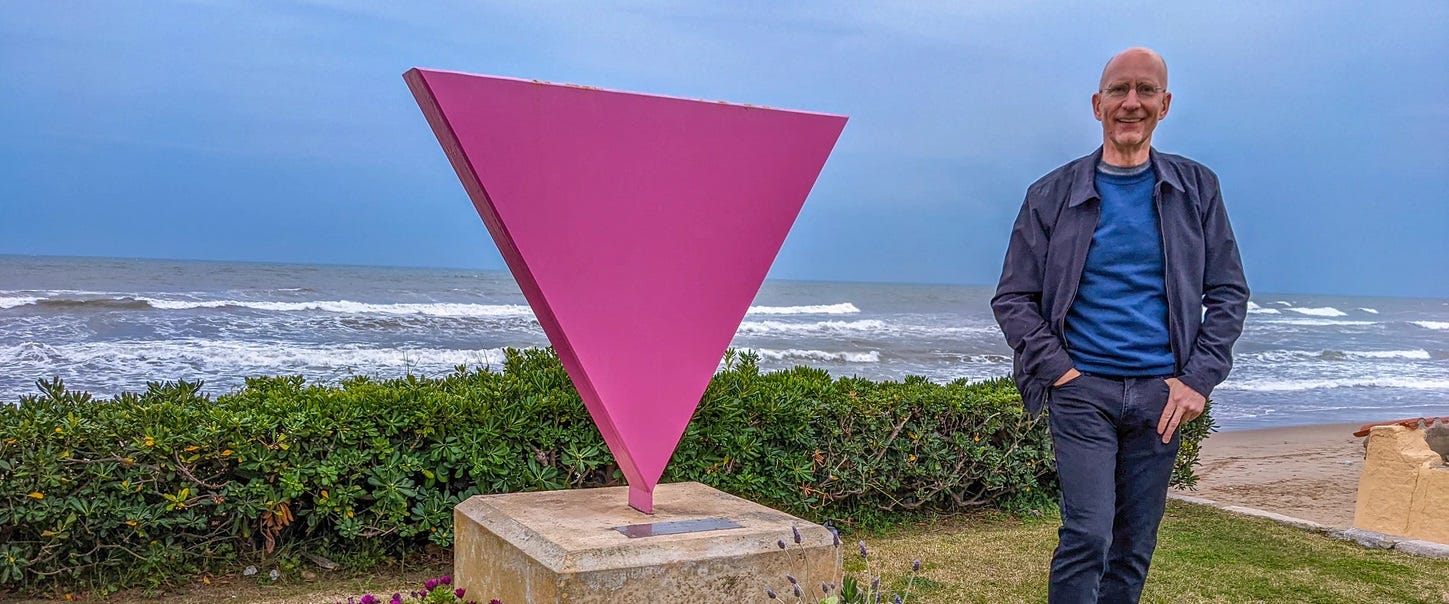 Brent standing next to a sculpture of the Pink Triangle LGBTQ Pride movement on the boardwalk in Sitges. 