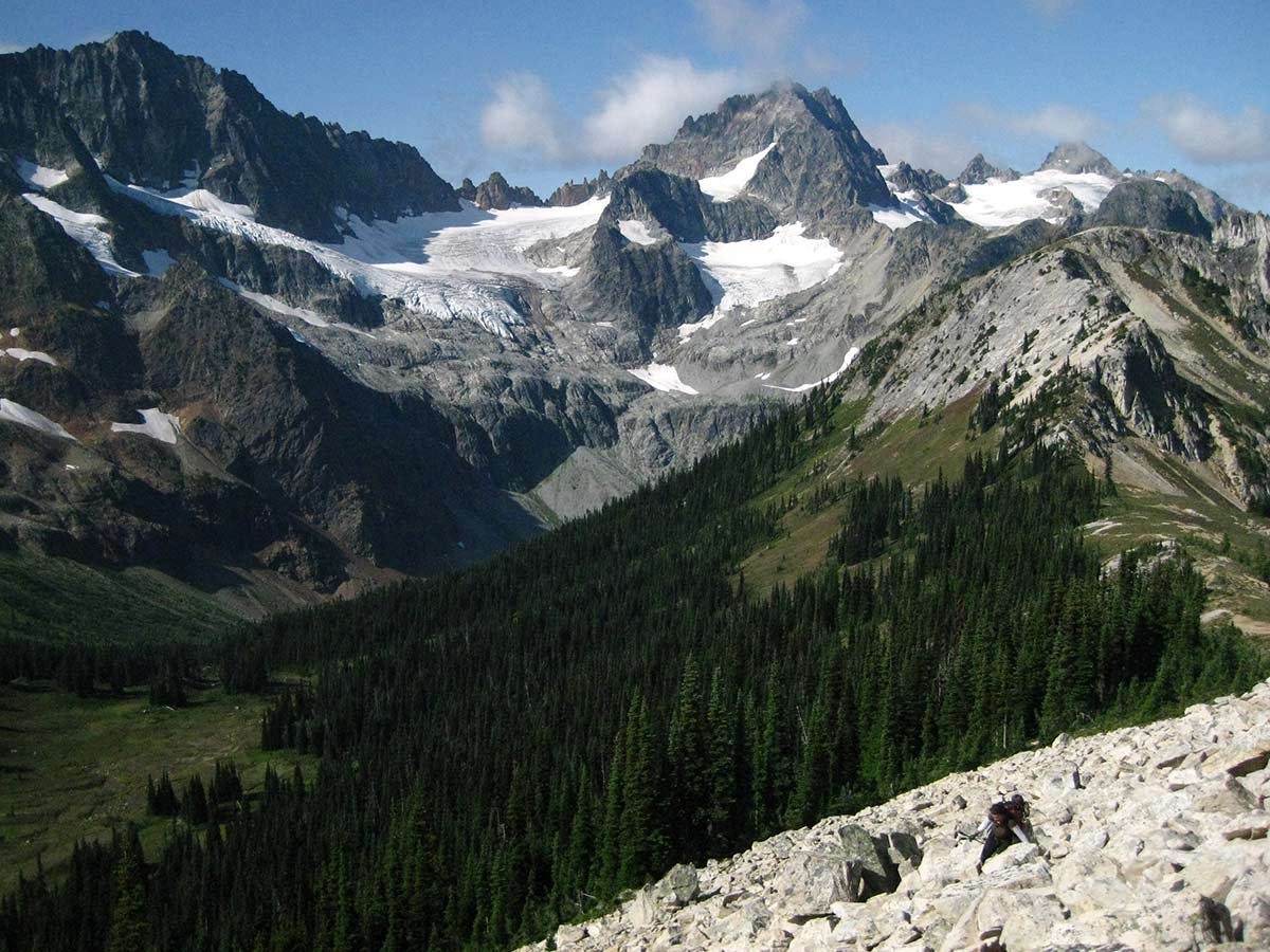 a woman in a large backpack holding trekking poles clambers over white boulders before an arcing ridgeline that sweeps down a forested slope to an open meadow below, snow-spotted Kitling Peak behind
