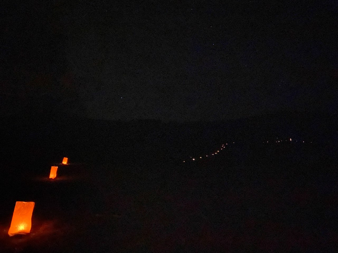 A black hillside at night with three luminarias in the foreground and numerous snaking off into the darkness over the hill in the background.