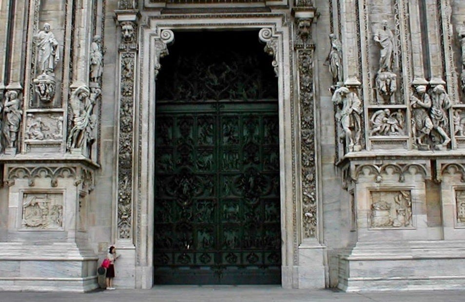 The massive carved steel doors of the Duomo in Milan Italy; a woman stands to the left of the doors illustrating their size in comparison to her