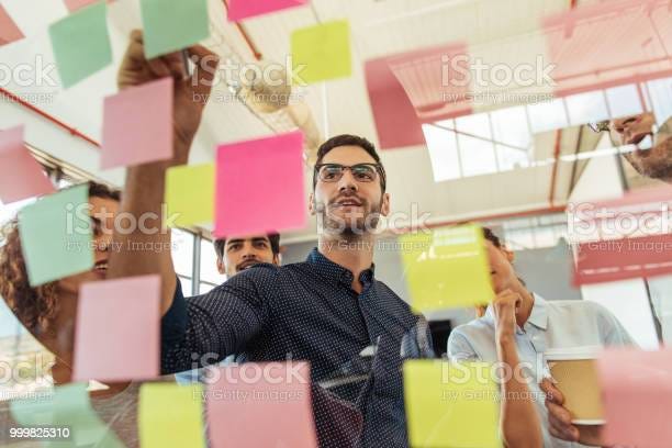 A group of people smiling and interacting with sticky notes on a glass wall with iStock watermark