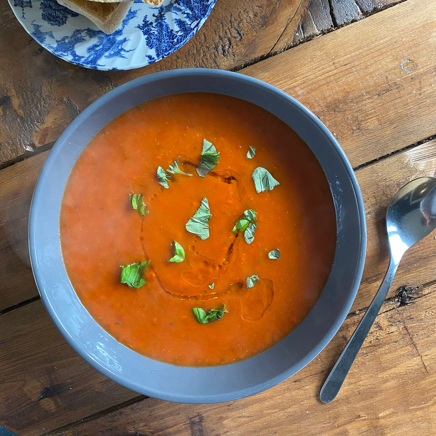 Grey bowl filled with tomato and pepper soup, topped with fresh basil. A small blue and white plate is to the side with a piece of toasted ciabatta bread. 