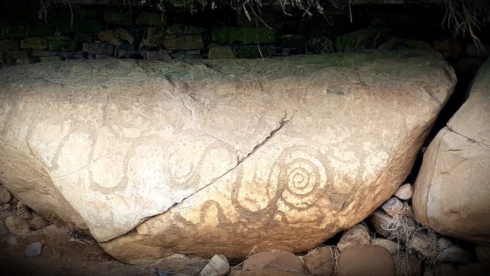 Large oval shaped kerbstone at Knowth, sandy coloured, large crack running top right to bottom left, with series of undulating wavy lines across surface and one prominent circular swirling motif right of centre.