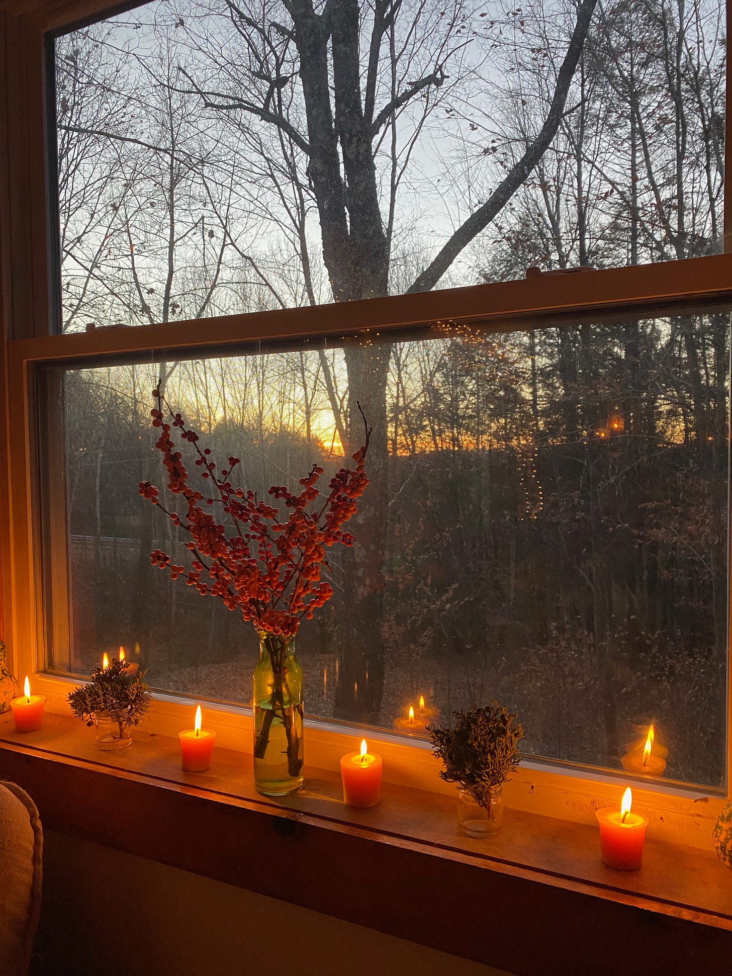 Four small beeswax candles on a windowsill. A large green vase of winterberry and two small glass jars of cedar sit between the candles. The view outside the window is dusky, with bare trees and a golden sun setting on the horizon.