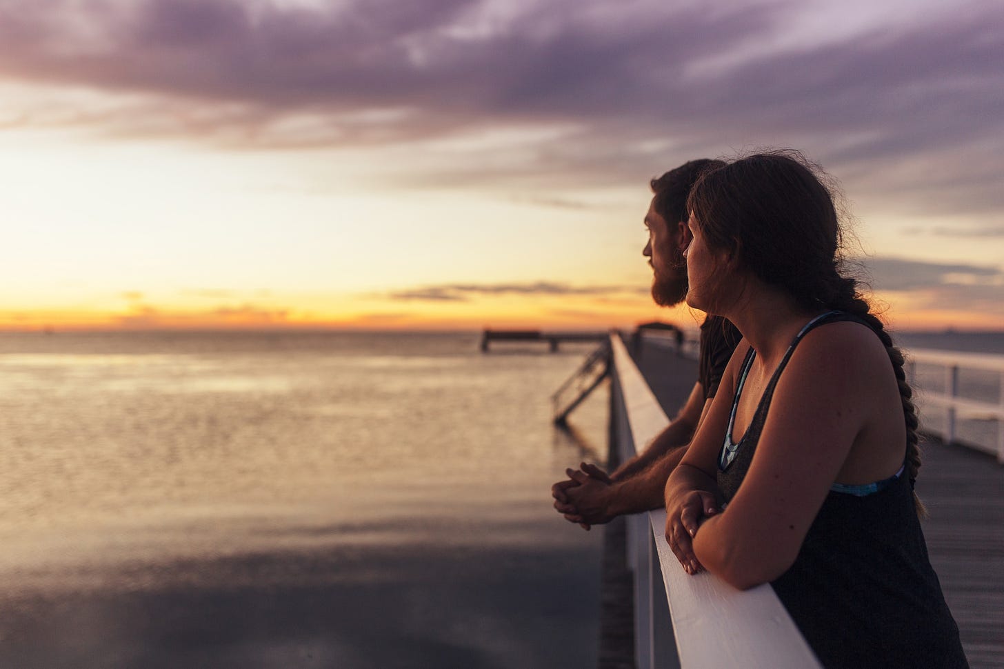 A pensive couple stands on a pier and look at the sunset. They wonder if their relationship will save them.