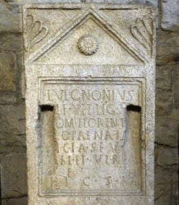 a photo of a roman tombstone with a triangular pediment at the top flanked by floral designs enclosing a circular motif. Underneath the pediment is an inscription field with latin letters. The inscription is damaged with two deep rectangular grooves flanking the inscription field