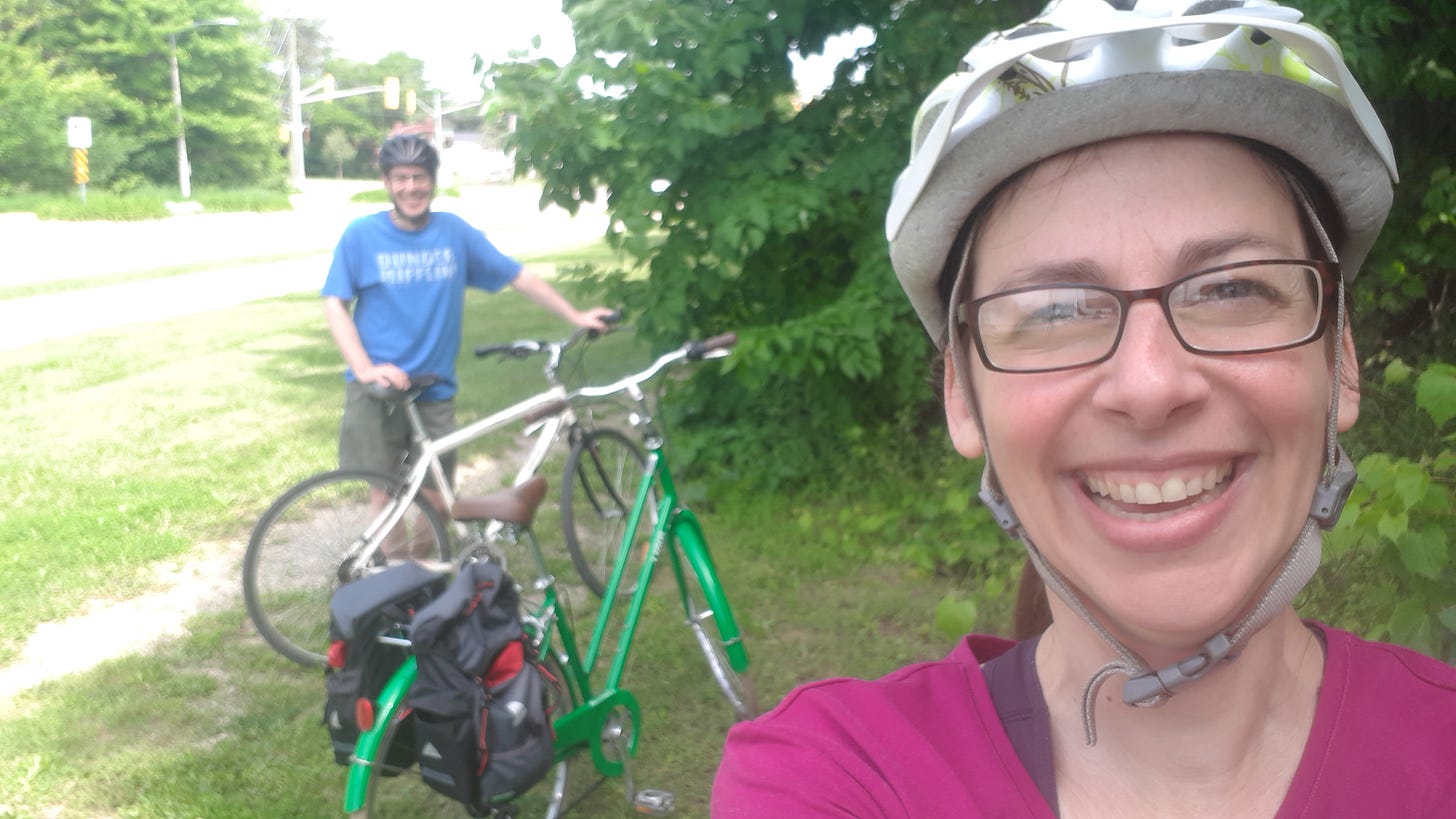 Woman (me) smiling, wearing a bike helmet. In the background there are 2 bikes and a man smiling.