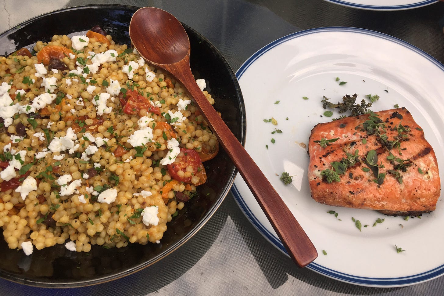 A black shallow bowl of pearl couscous salad with roasted tomatoes and olives, with herbs and feta on top. Next to is is a white plate with a deep pink salmon fillet, covered with a dusting of pepper and some fresh marjoram.