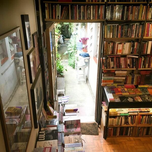 Foto tirada de um lugar elevado. No centro, uma porta aberta dando saída para um corredor iluminado. Plantas estão nesse corredor. Na parte de dentro, uma livraria. Os livros permeiam toda a parede da porta, inclusive na parte de cima do batente. Na outra parede da foto, quadros não-identificados estão pendurados e uma mesa na parte de baixo expõe diversos livros.