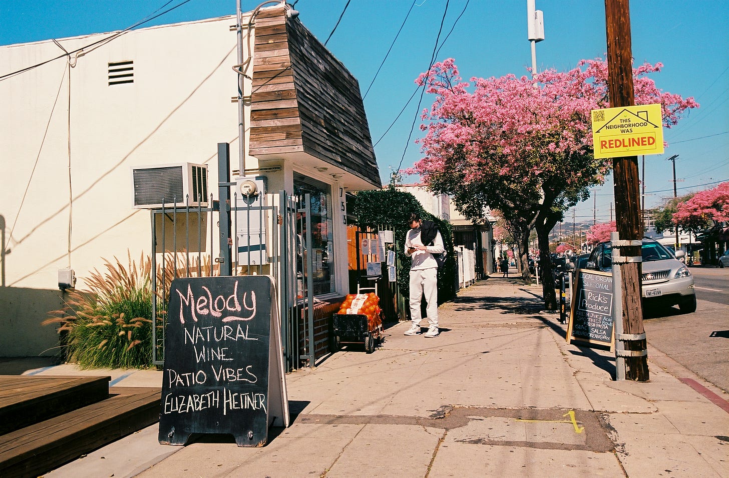 Photograph of Melody Wine Bar on the left side of the frame and one of my signs nailed to a wooden pole on the left side of the frame with the pink trumpet tree lined Virgil Ave in the background.