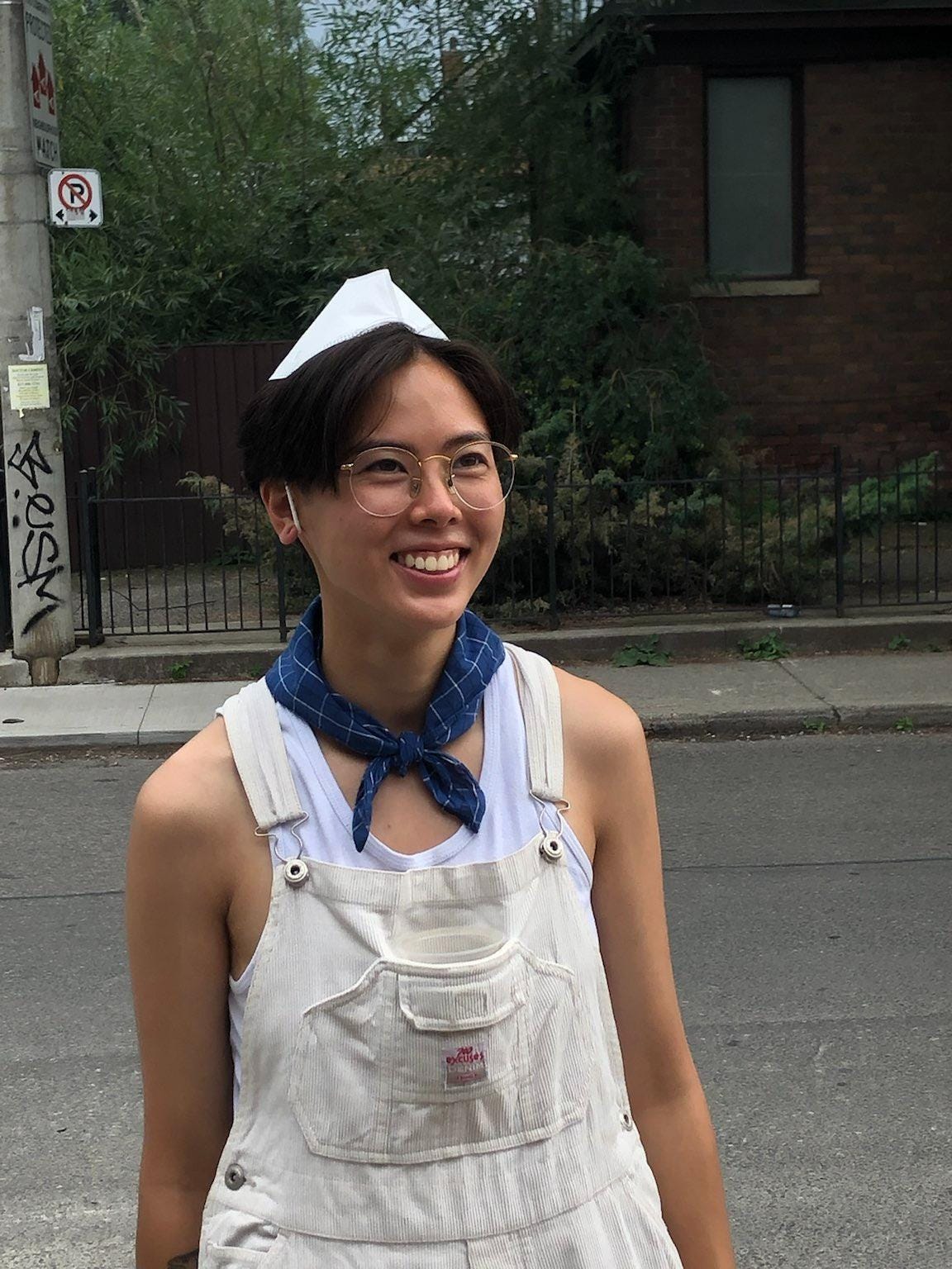 Khanh standing on the street in white overalls, and a blue bandana around their neck. They are wearing a KN95 mask on their head and smiling to their friend just off camera