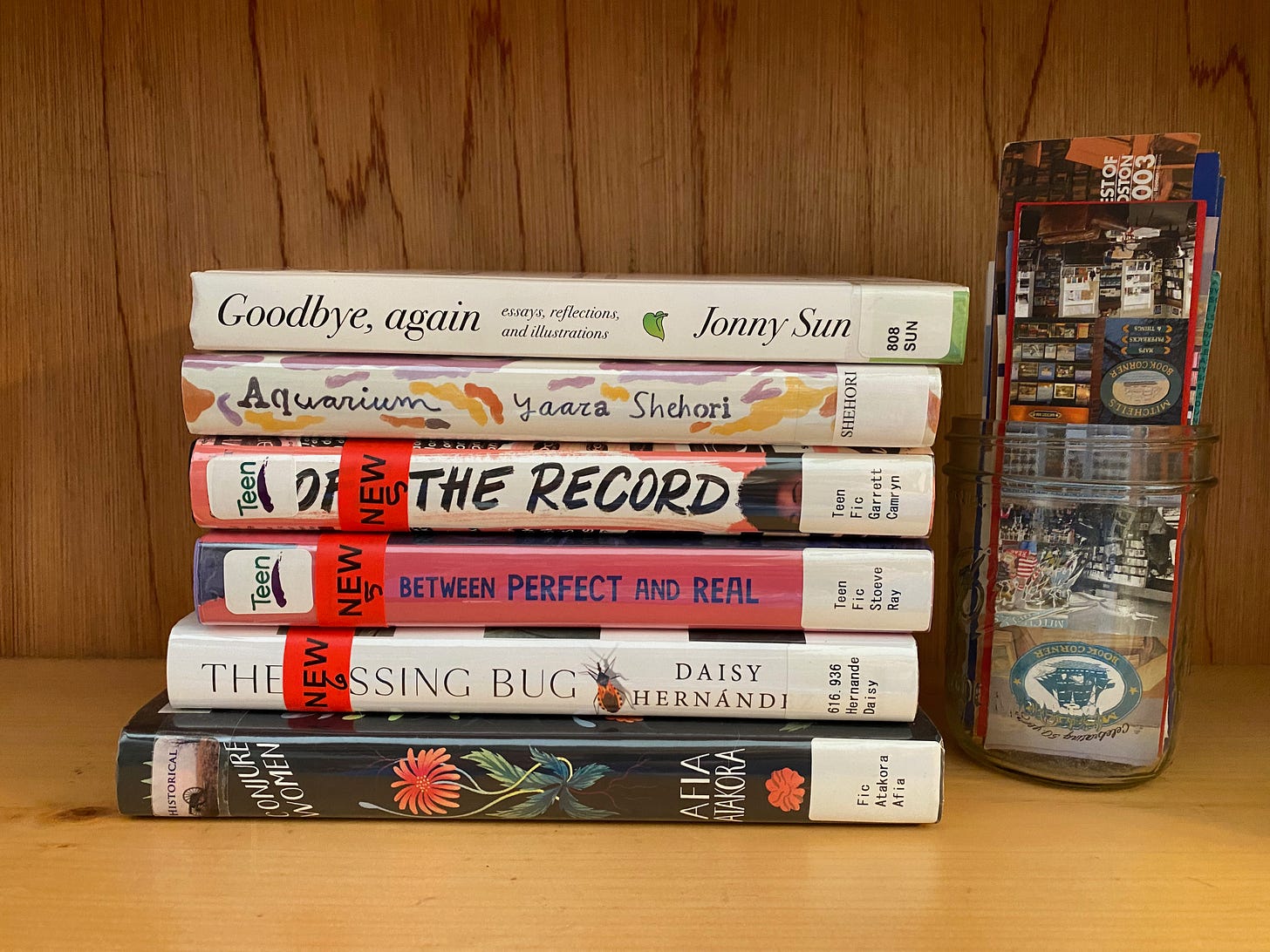 Closeup of a  stack of library books on a wooden shelf, sitting next to a jar of bookmarks. The books are: Conjure Women, The Kissing Bug, Between Perfect and Real, Off the Record, Aquarium, and Goodbye, Again.