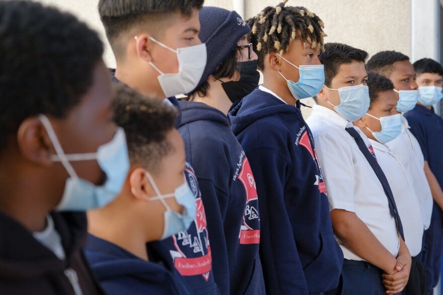 Adolescent boys, stand in a line with face masks, school sweatshirts or white button-downs and ties. They are shown from the side. 