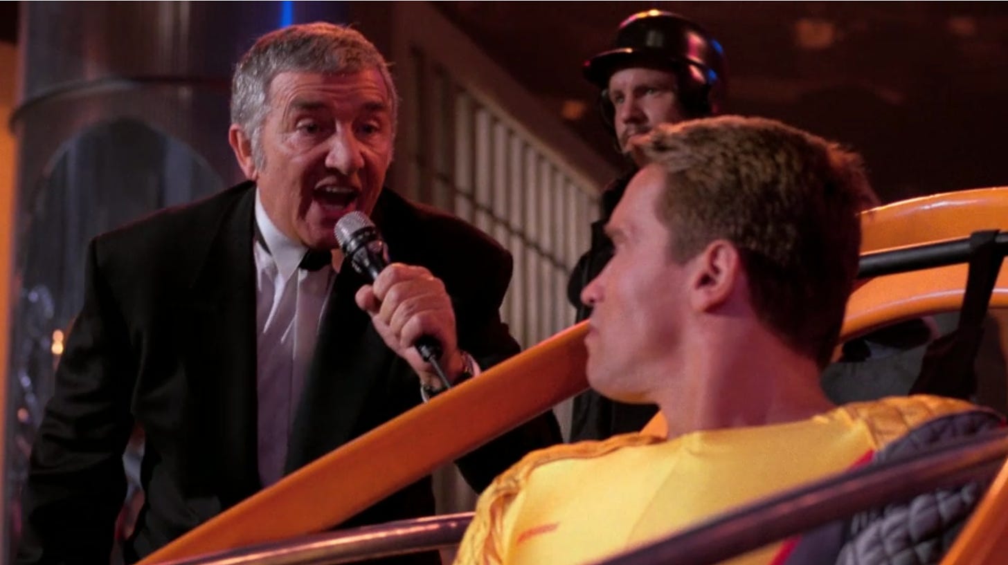 Richard Dawson, holding a microphone, addresses Arnold Schwarzenegger as Schwarzenegger, wearing a yellow spandex suit, glares at him from the cockpit of an orange-framed rocket sled. 