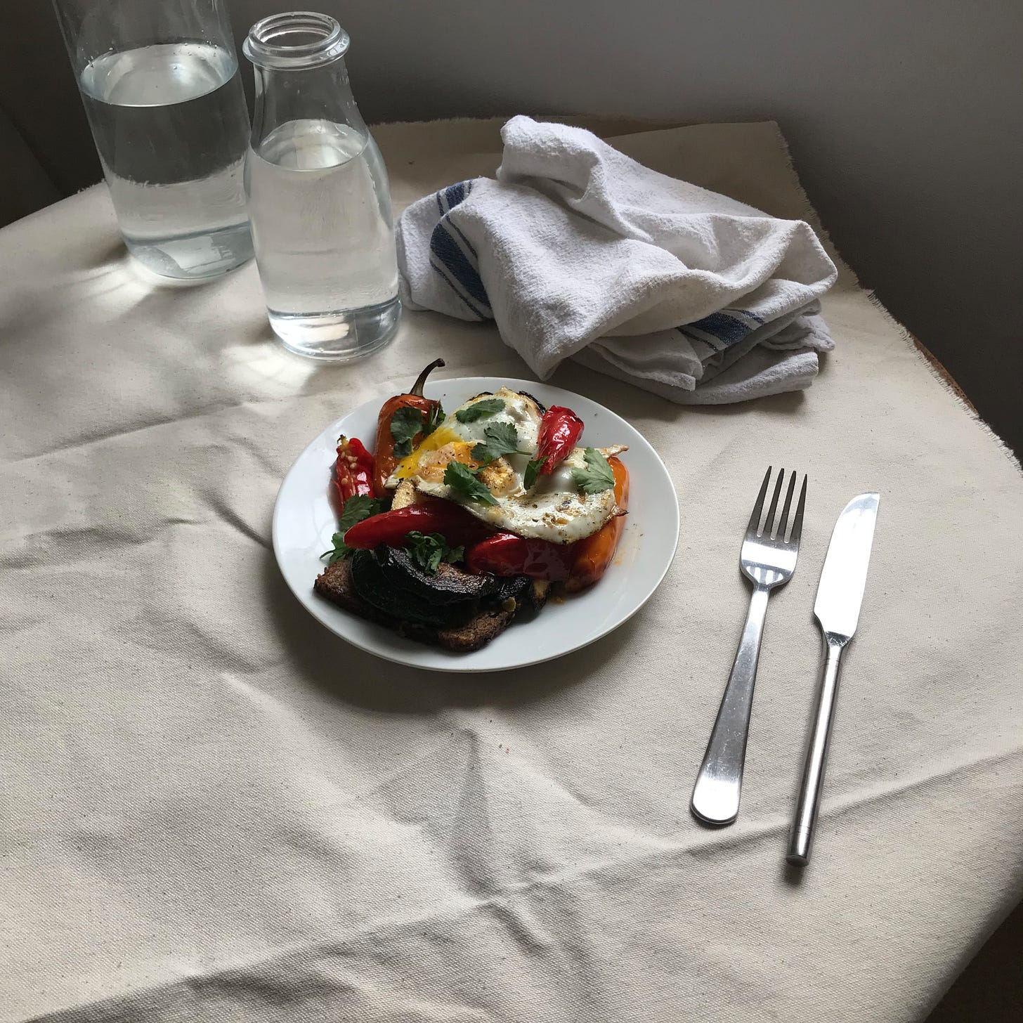 A white plate of food set on a table with cutlery, a jumbled kitchen towel, two glass carafe of water. On the plate, in the centre, mounded up, some dark brown bread topped with soft fried eggs and some small long red peppers, a green herb too. 