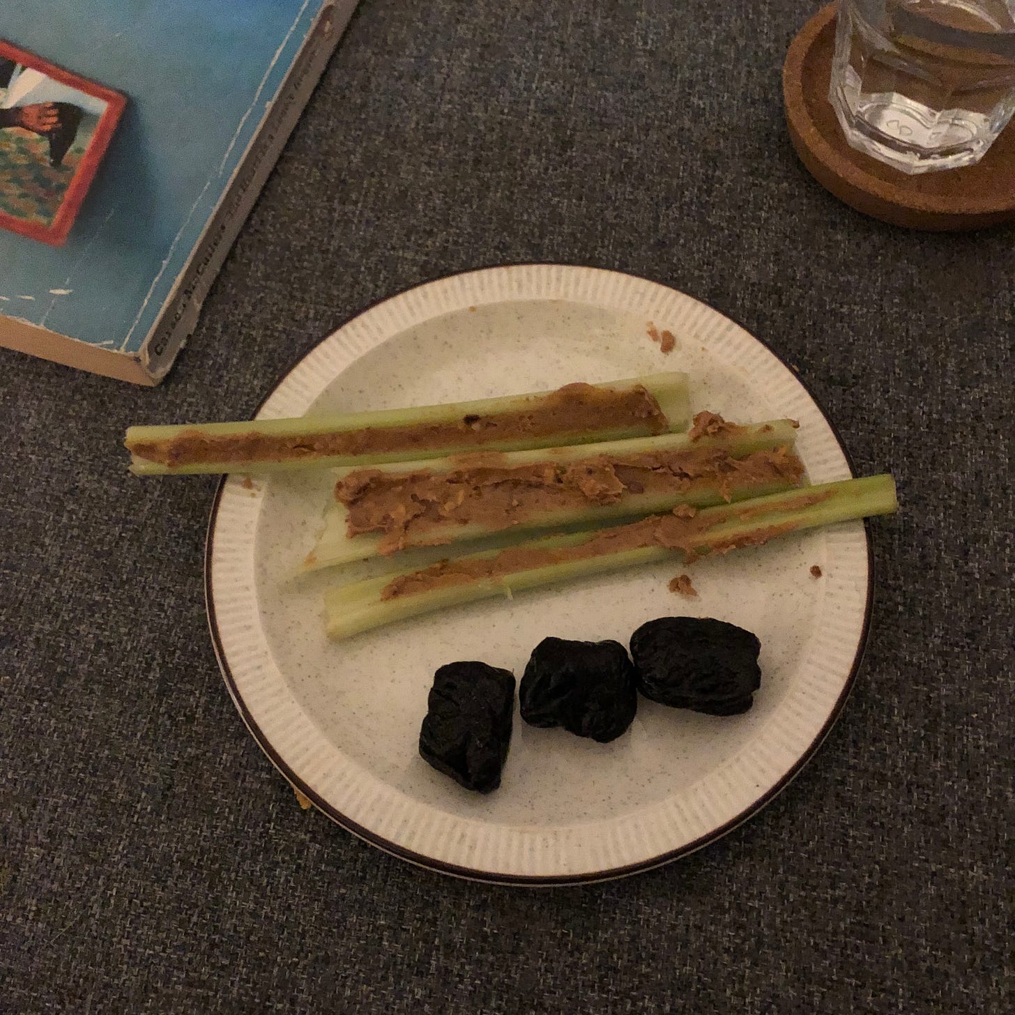 A small plate with three lengths of celery, with peanut butter spread into their channels, and three dark wrinkly prunes. Beside this, the edge of a book and a glass of water on a cork coaster. 