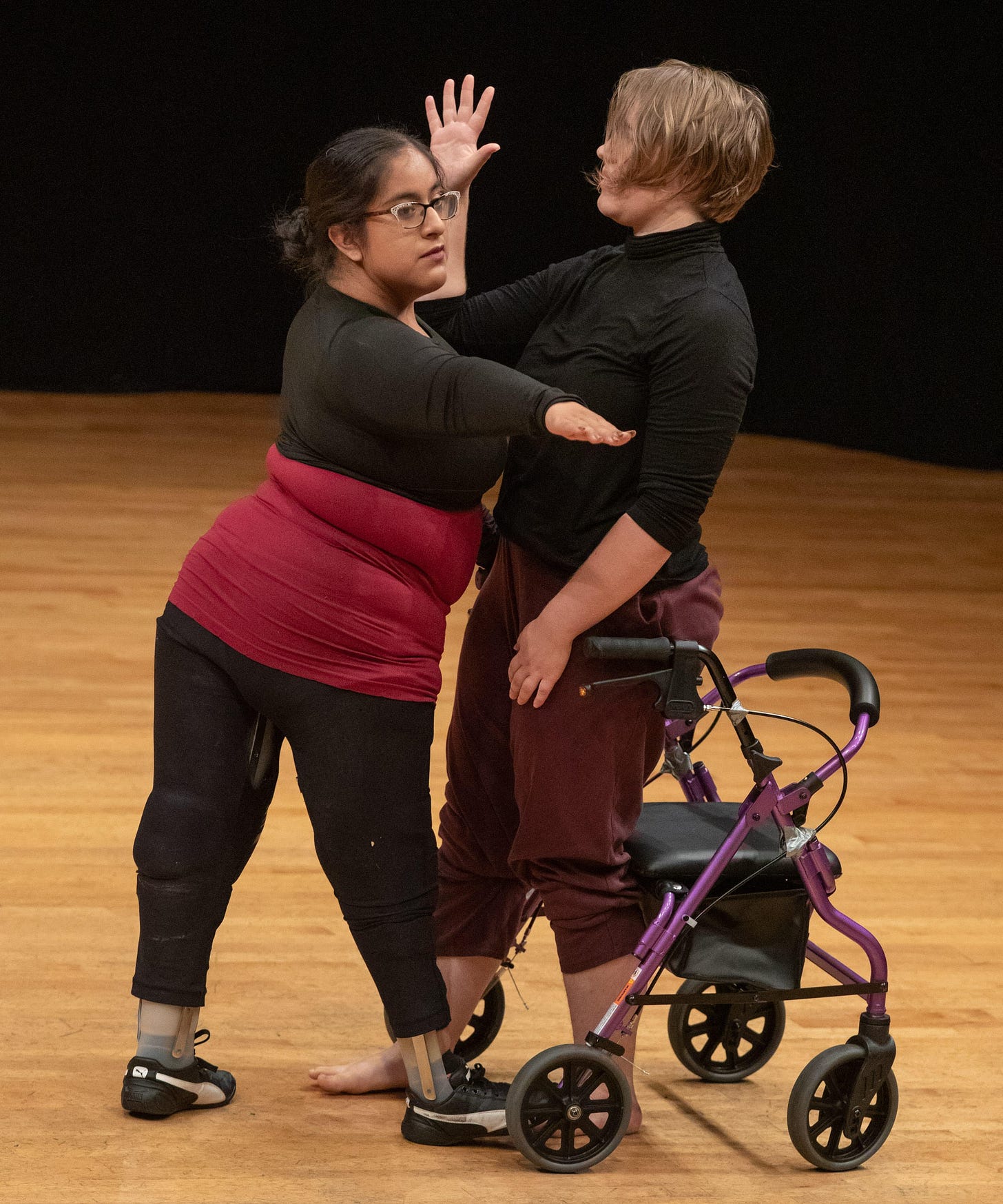 Two disabled dancers stand close, one’s gaze following an outstretched hand, the other facing her with a palm straight up. One dancer wears leg braces, the other has a wheeled walker/chair nearby.