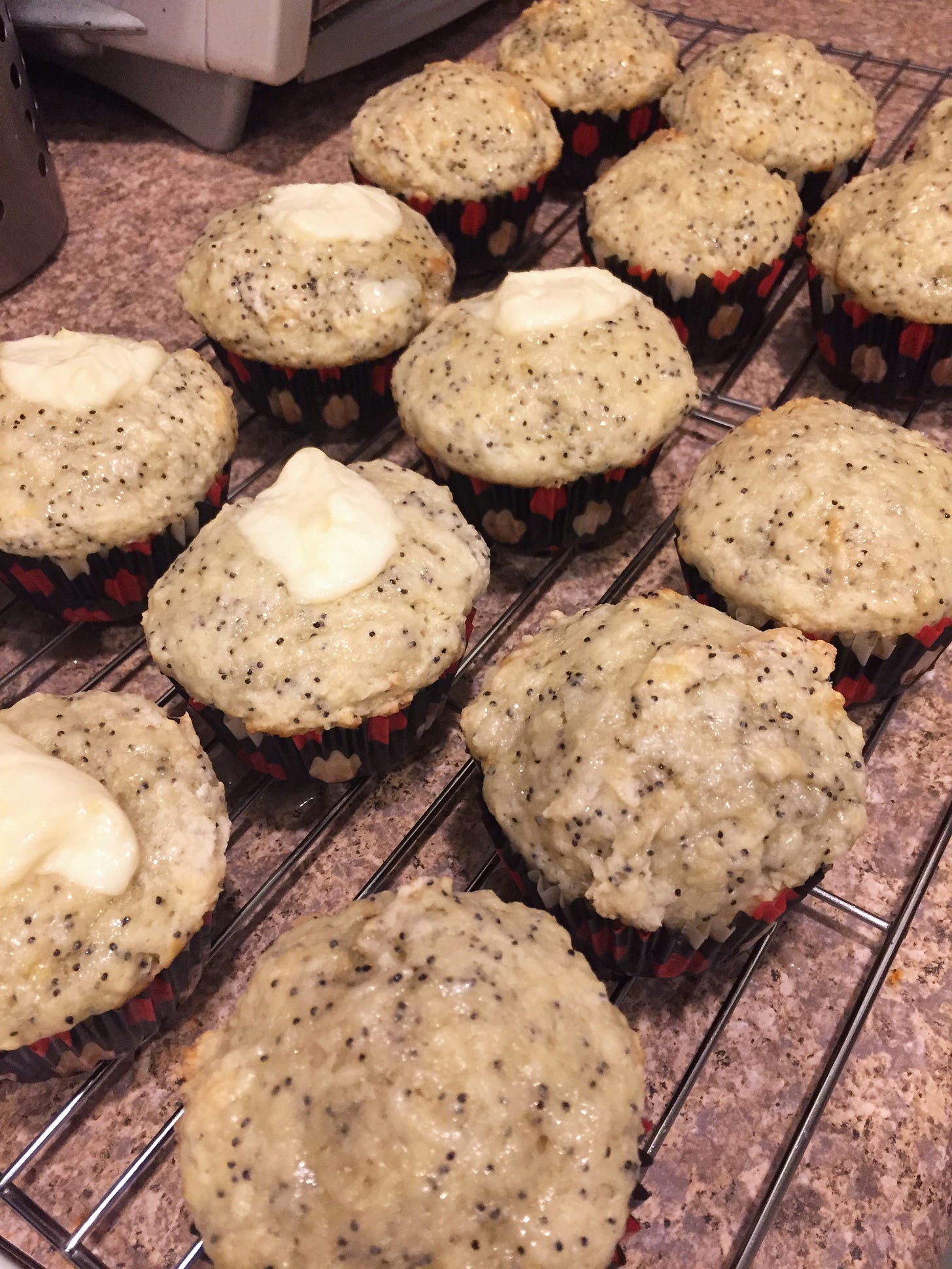 Lemon muffins, some in the foreground with blobs of mascarpone on top, dotted with poppyseeds sit on a cooling rack.