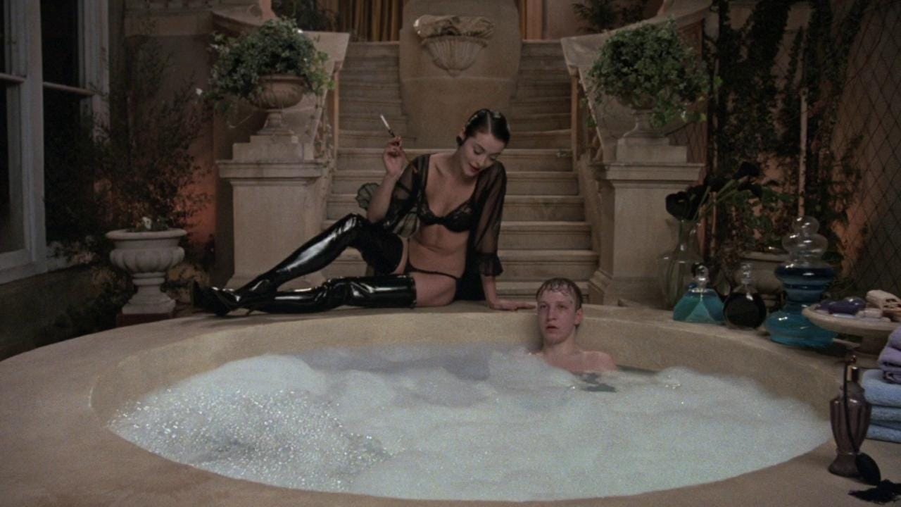 Amanda Donohoe sitting and smoking on the edge of a bathtub in lingerie, about to kill an unsuspecting young man, in Ken Russell’s "The Lair of the White Worm" (1988)