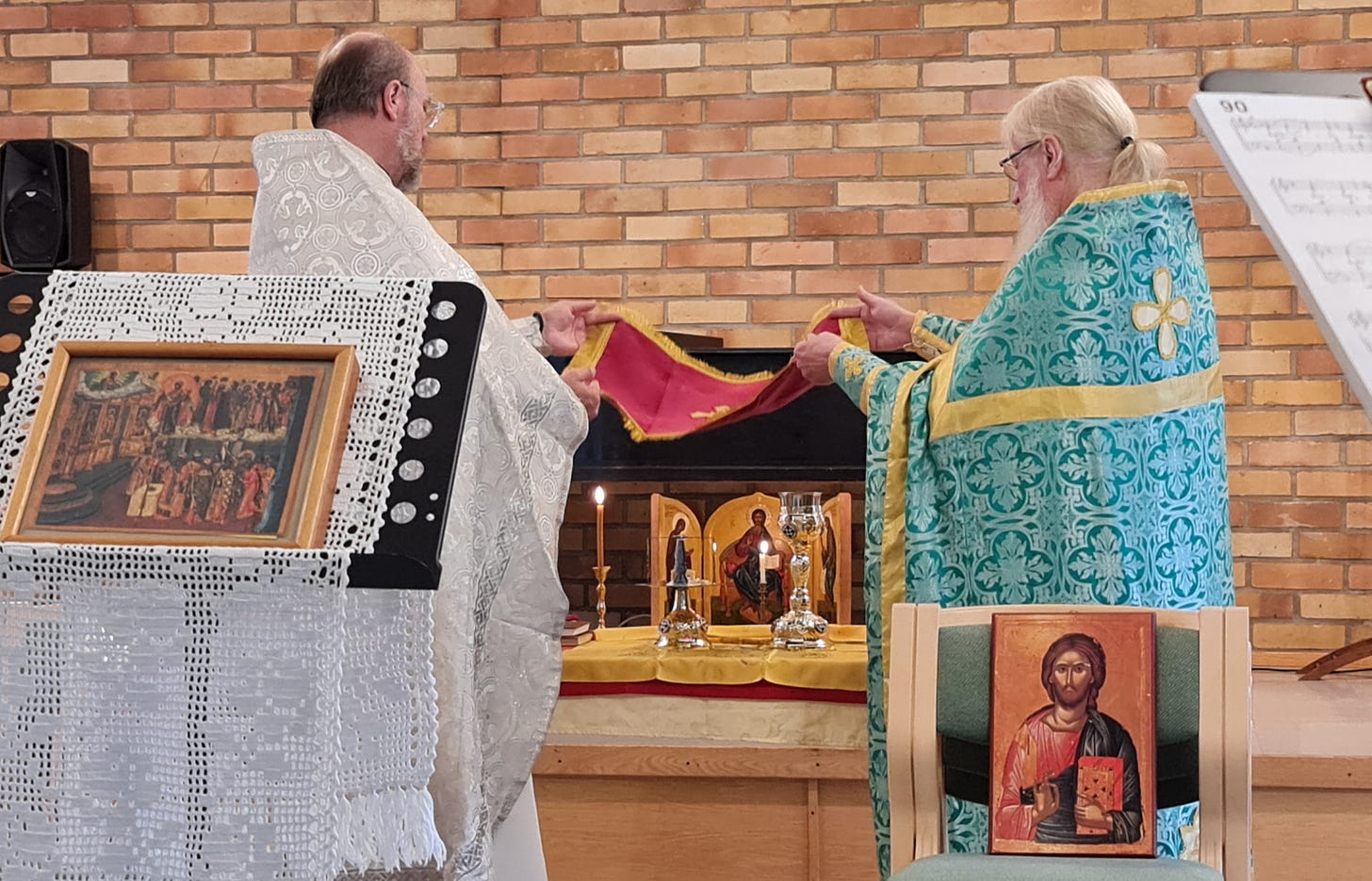 Två ortodoxa präster står vid ett altarbord och håller i en röd duk med ett kors på över de eukaristiska gårvorna. I förgrunden syns två ikoner, den ena framställd på ett notställ och den andra på en stol.