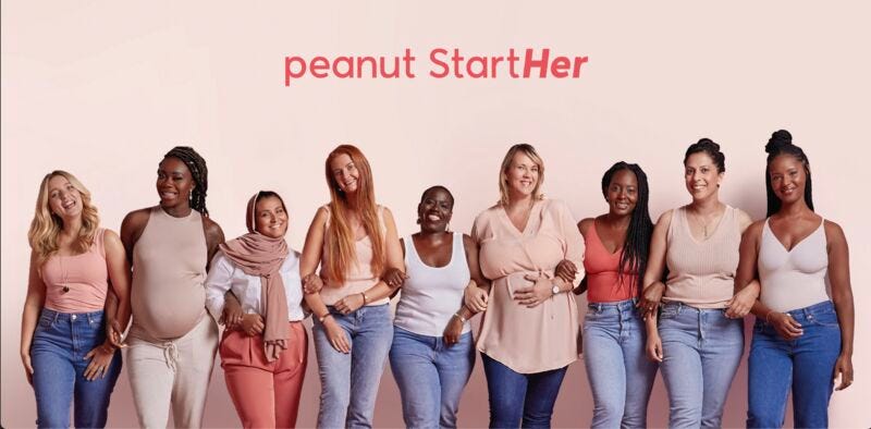 Nine women are standing side by side, interlocking their arms. They’re all smiling to the camera.Most of them are wearing blue jeans and nude coloured tops. Above the women, a coral-coloured text says: peanut Start Her, with the last two words attached to one another.