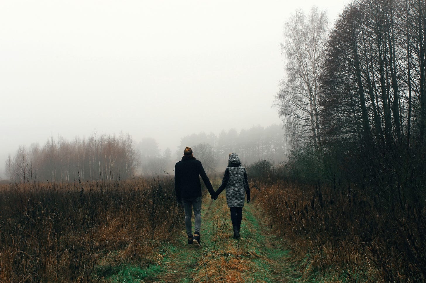 A couple holds hands and walks down a foggy country path lined with trees and fields.