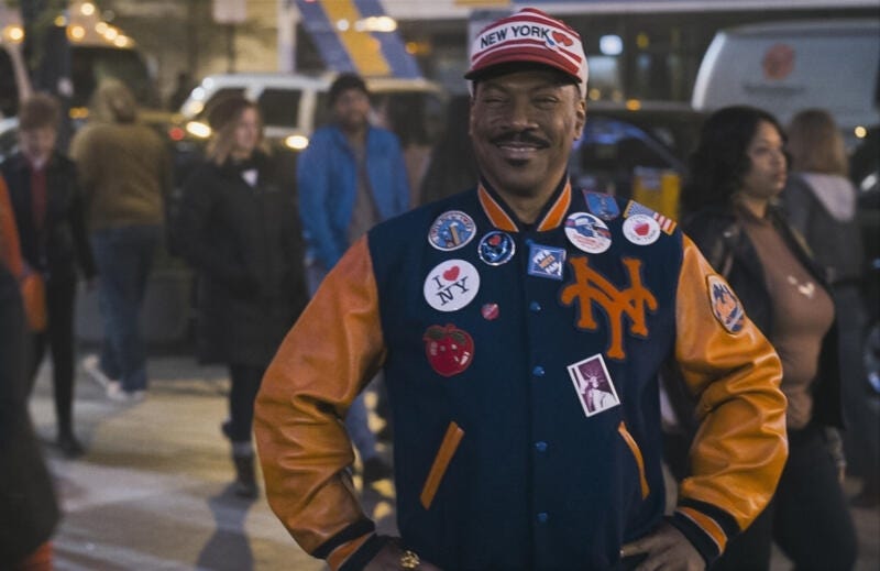Eddie Murphy as King Akeem in 'Coming 2 America', smiling towards the camera, in the middle of a street crowd, wearing New York iconography.