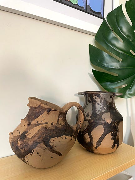 Two ceramic pots on a shelf with a monstera leave in background.