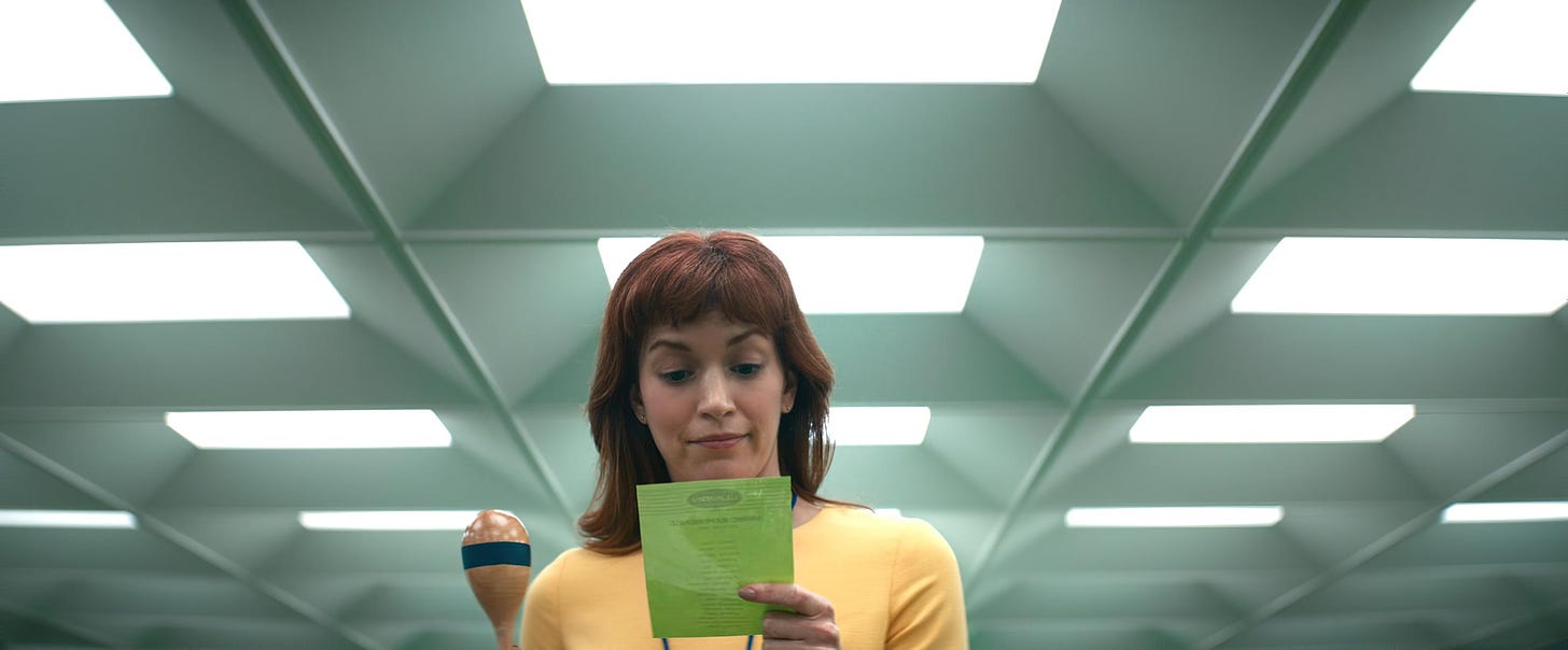 Low shot of an auburn-haired woman in a yellow top under pitiless fluorescent office lights, looking down at a green card and holding a maraca in her right hand