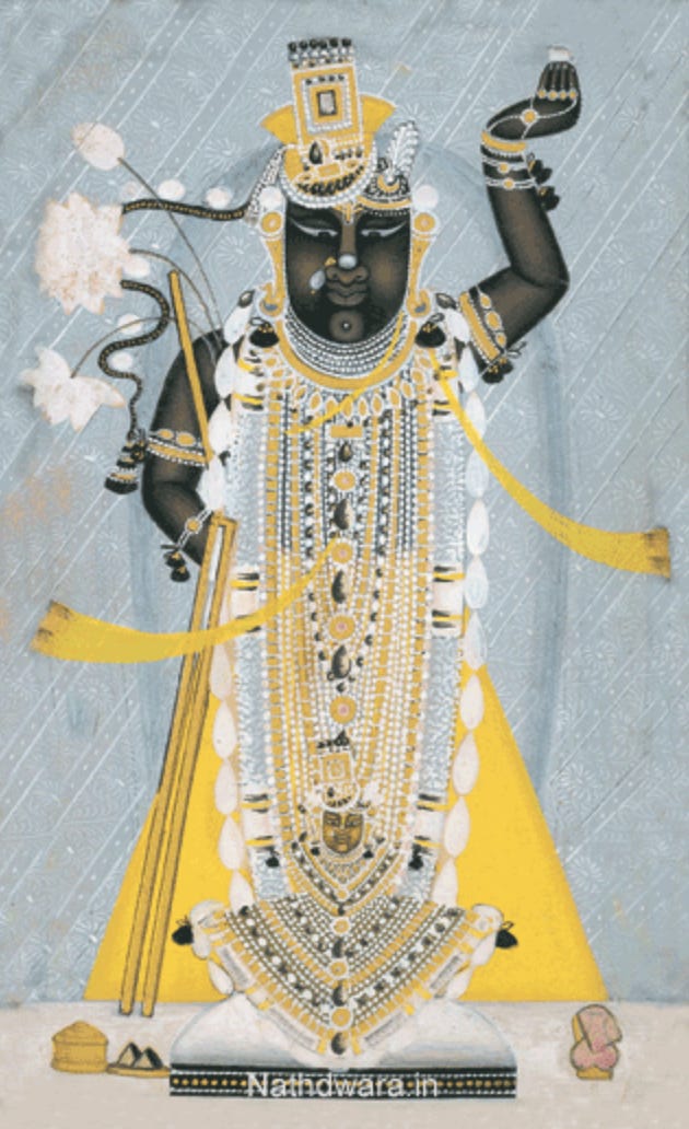 A black deity who stands with left arm raised up and fingers pointing upwards. The deity is wearing yellow robes, with many jewel necklaces, bracelets, earrings, and crown.