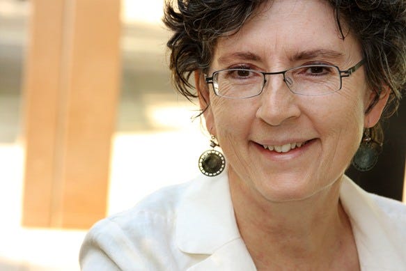 A white woman with short curly dark hair and rectangular glasses and circular earrings smiles, looking at the camera.