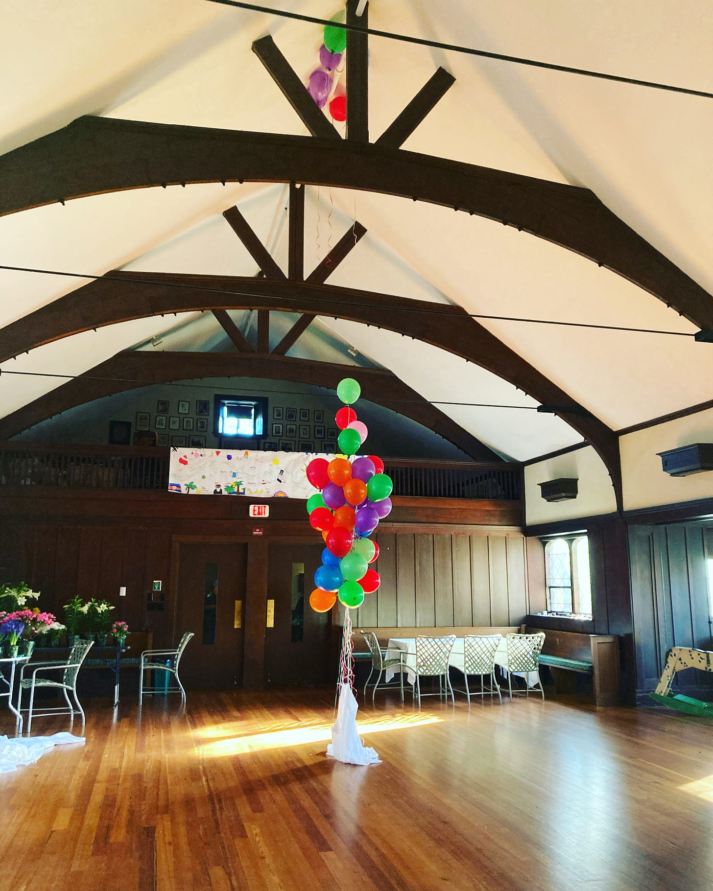 A large bundle of multicolored balloons float in the middle of a big hall with a white sheet hanging from the bottom of the bundle.