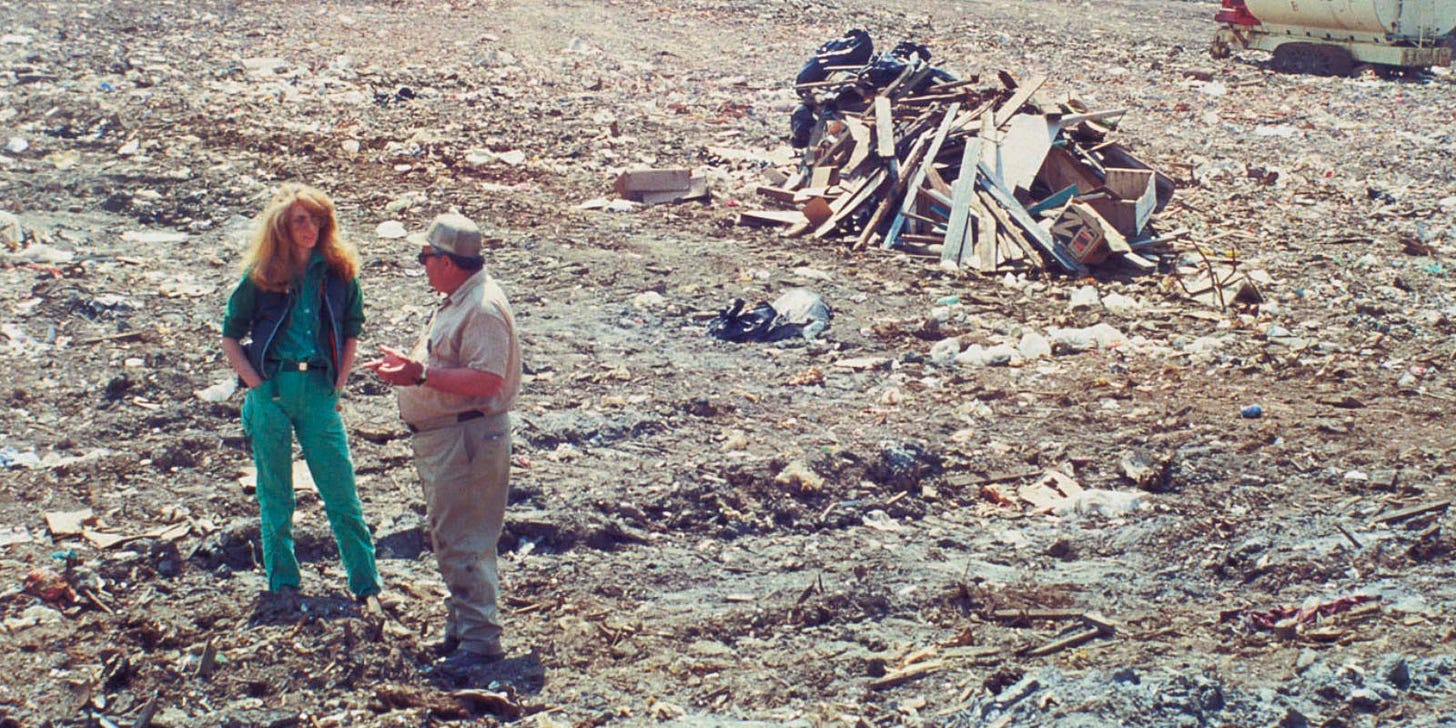 Photo of a woman, hand in her pockets, having a conversation with a man. Both people are at a dump site. The woman is Mierle Laderman Ukeles. In the background of the photo, is a pile of garbage. The man is a sanitation worker. Both persons are standing on a landfill in the sun.
