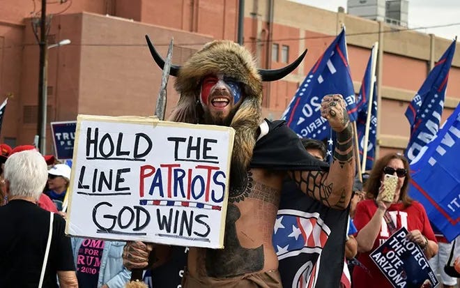 Photo of QAnon Shaman on January 6 holding sign that says "Hold the line patriots, God wins"