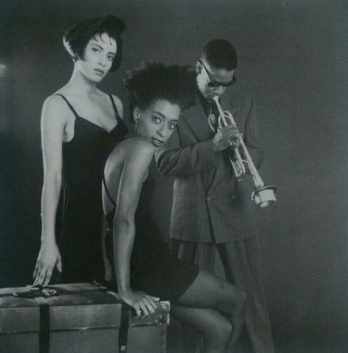 This production still from Spike Lee's 1990 film Mo' Better Blues shows the characters Clarke Betancourt (played by Cynda Williams, left), Indigo Downes (played by Joie Lee, center), and Bleek Williams (played by Denzel Washington, right) arrayed near a steamer trunk. Clarke and Indigo, wearing skimpy black dresses, look directly into the camera, while Bleek, wearing a full suit and sunglasses, plays a muted trumpet pointed toward the floor. This production still from Spike Lee's 1990 film Mo' Better Blues shows the characters Clarke Betancourt (played by Cynda Williams, left), Indigo Downes (played by Joie Lee, center), and Bleek Williams (played by Denzel Washington, right) arrayed near a steamer trunk. Clarke and Indigo, wearing skimpy black dresses, look directly into the camera, while Bleek, wearing a full suit and sunglasses, plays a muted trumpet pointed toward the floor.