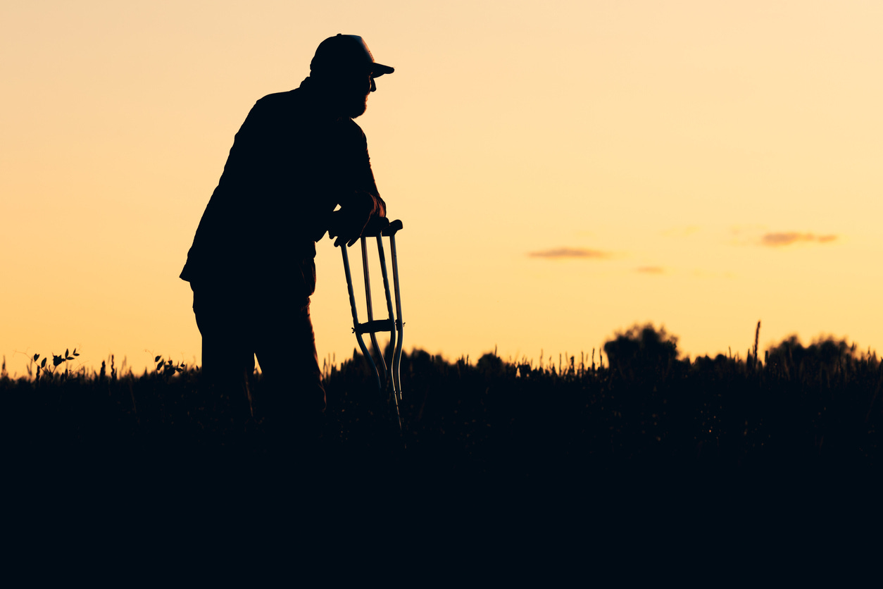 Silhouette of older man with cap leaning on his pair of crutches against sunset sky.