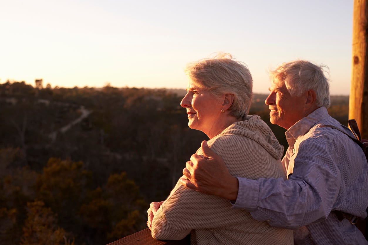 Gray-haired caucasion older couple looking out of deck at sunset. The man has his left hand on his wife's shoulder. Sunset provides profile of each person's attractive face.