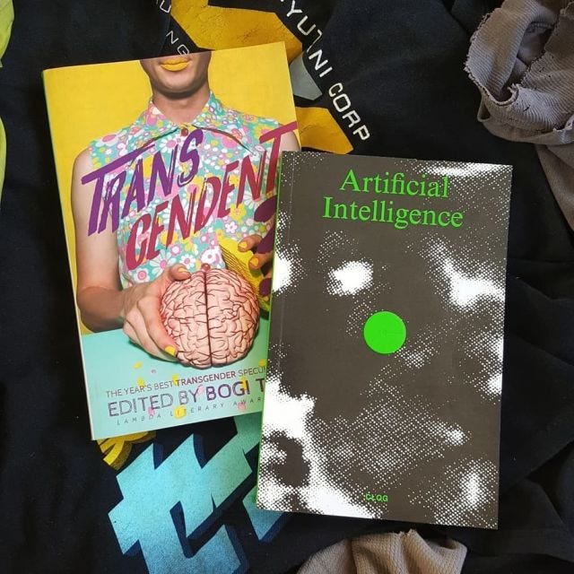 Transcendent 3 & CLOG MAgazine's ARTIFICIAL INTELLIGENCE issue lay on top of a pile of black t-shirts